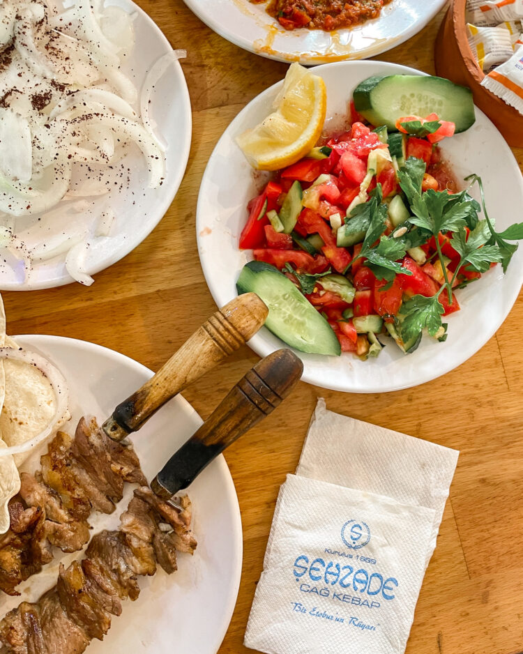 Cag kebab on a wooden skewer, plate of onions sprinkled with sumac and a chopped tomato salad.